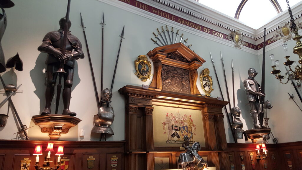 Interior view of a medieval great hall featuring full suits of plate armor on wall brackets, spears, shields, and detailed wood paneling. The text "THE WHOLE ARMOUR OF GOD" is visible on the upper wall border.