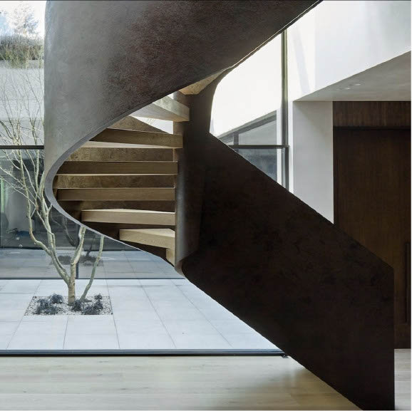 Close-up of a modern, minimalist cantilevered spiral staircase. Features floating light wood treads contrasted against a dark, textured corten steel or blackened bronze inner spine and handrail, adjacent to a large glass window wall.